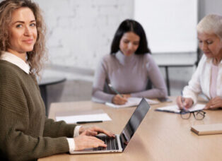 A woman with curly hair sits at a table, typing on her laptop about the latest online 3 Phase UPS, and smiling. Two other women in the background are focused on writing notes. The setting appears to be a meeting or collaborative work environment.