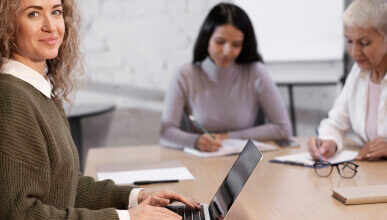 A woman with curly hair sits at a table, typing on her laptop about the latest online 3 Phase UPS, and smiling. Two other women in the background are focused on writing notes. The setting appears to be a meeting or collaborative work environment.