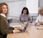 A woman with curly hair sits at a table, typing on her laptop about the latest online 3 Phase UPS, and smiling. Two other women in the background are focused on writing notes. The setting appears to be a meeting or collaborative work environment.