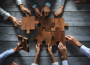 A group of hands assembles large wooden puzzle pieces on a rustic table, symbolizing teamwork and collaboration—much like configuring an Online 3 Phase UPS system for seamless power solutions.