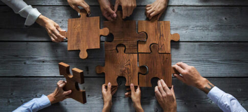 A group of hands assembles large wooden puzzle pieces on a rustic table, symbolizing teamwork and collaboration—much like configuring an Online 3 Phase UPS system for seamless power solutions.